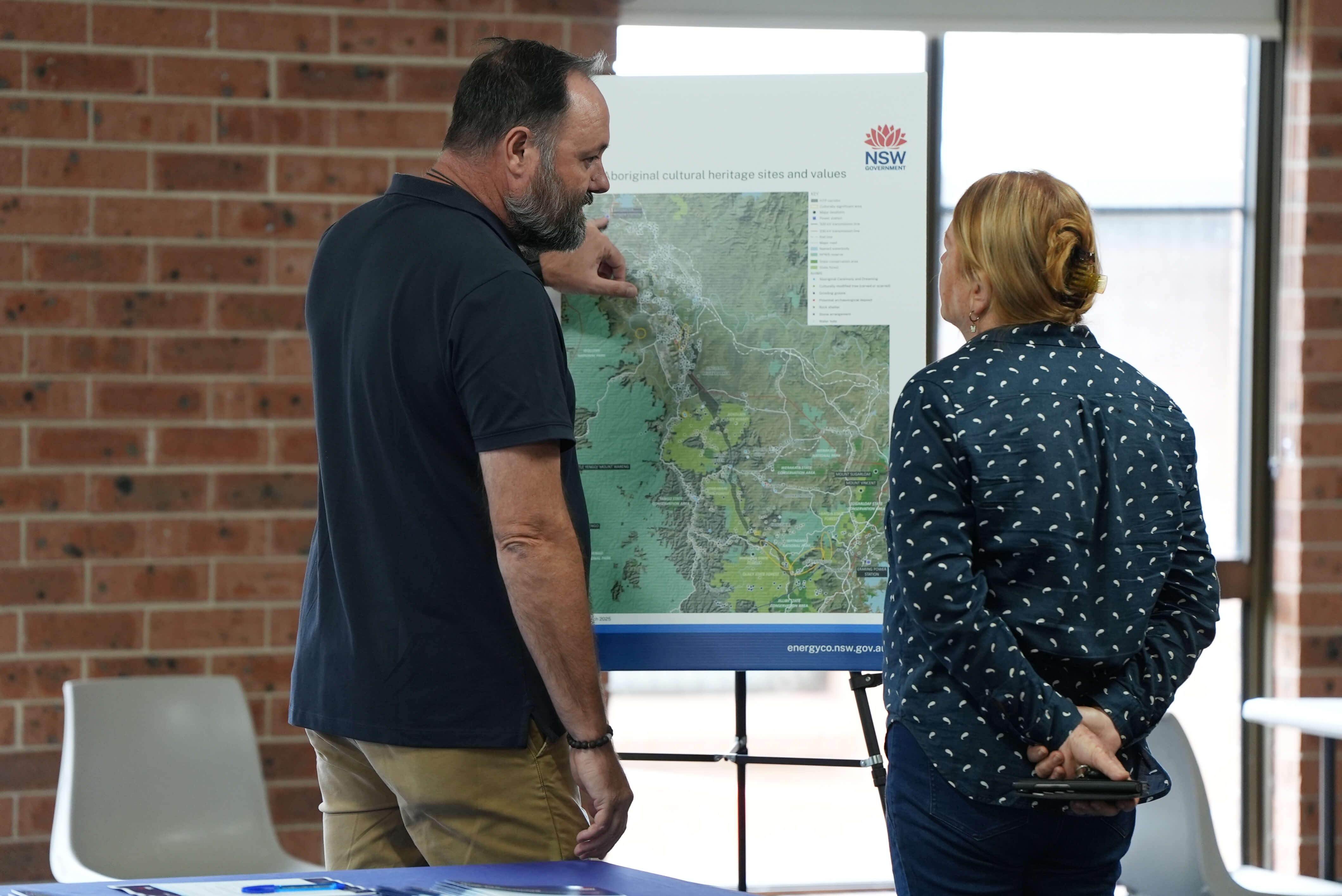 A man in a black polo and a woman in a navy and white shirt look at a map of the Hunter Transmission Project propped up on an easel.  