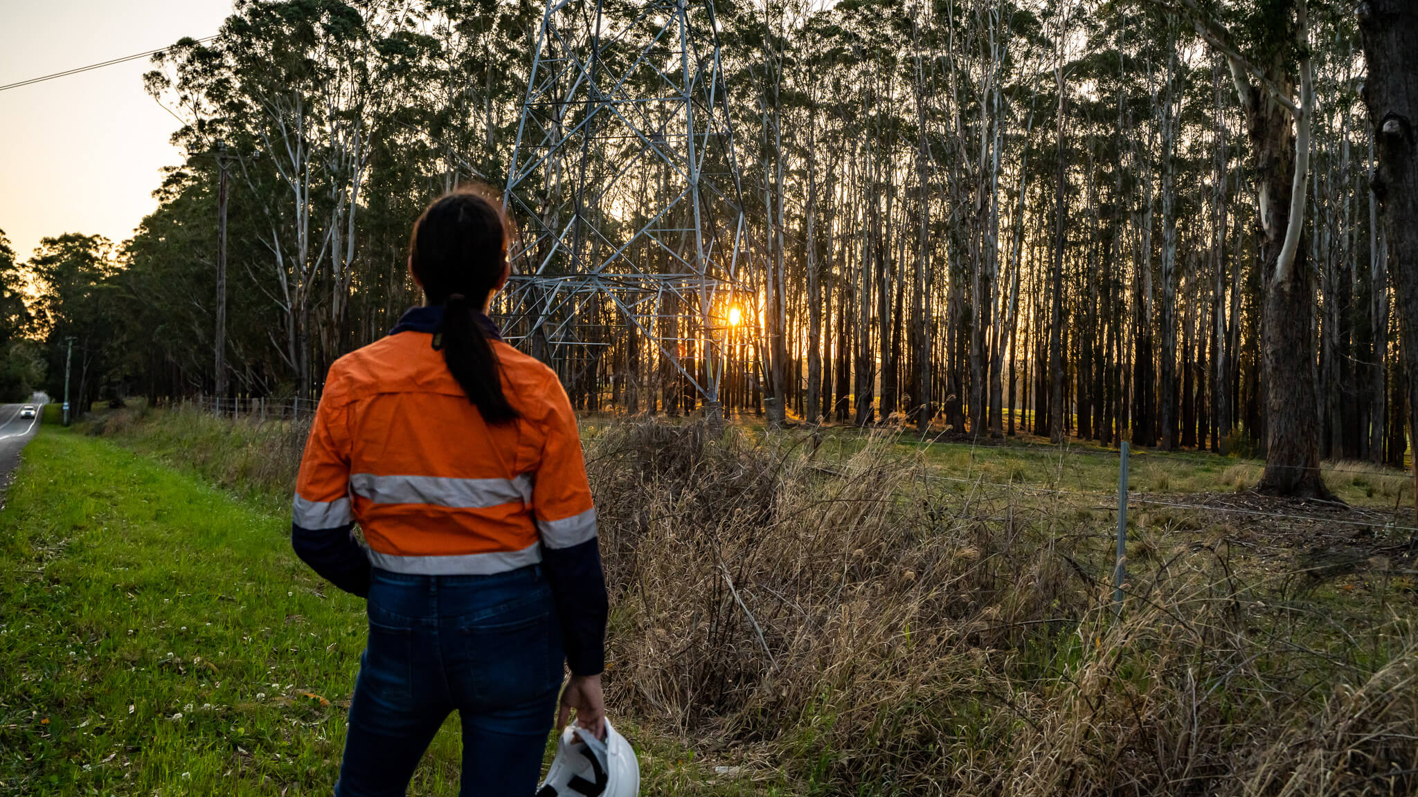Female worker in high vis jacket holding a hard hat looking at the base of the transmission lines